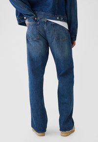Jean en denim bleu foncé avec une coupe droite. Il comporte deux poches arrière et une couture visible le long des jambes. Porté avec une veste en denim.