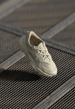 Beige sneaker with textured fabric, contrasting smooth accents, rounded sole, and laces, suspended above a gridded surface.