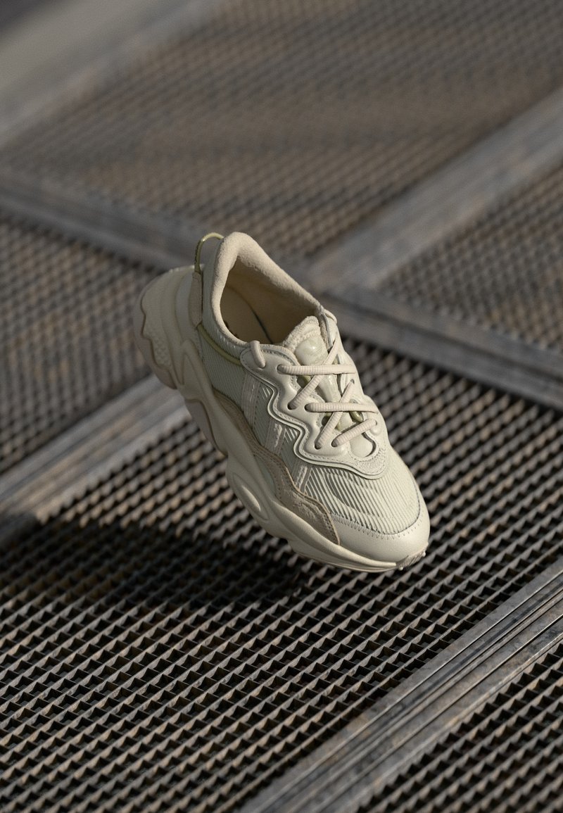 Sneaker beige con tessuto texturizzato, accenti lisci a contrasto, suola arrotondata e lacci, sospeso sopra una superficie a griglia.