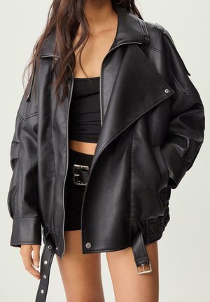 Vrouw draagt een oversized zwarte leren jas over een zwarte crop top en korte broek met een zilveren gesp riem, handen in de jaszakken.
