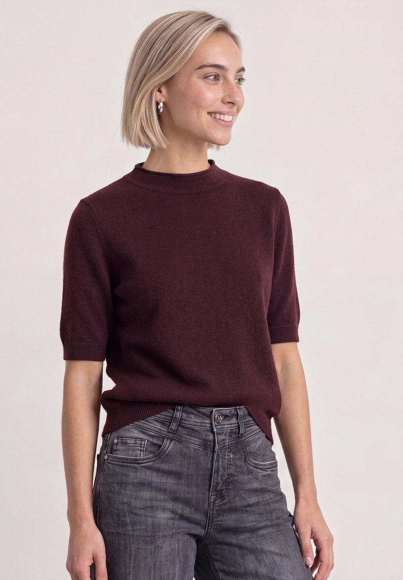Femme aux cheveux blonds courts portant un haut bordeaux à manches courtes et un jean gris, souriante et regardant vers la droite sur un fond uni.