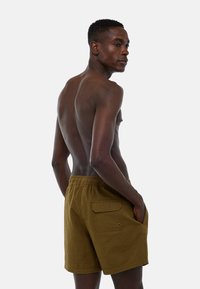 Shorts de bain bruns texturés avec une taille élastique et une poche latérale, présentés sur une personne vue de dos sur fond blanc.