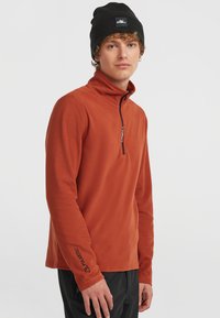 Orange fleece-tröja med halvdragkedja, med Polartec-logotyp på ärmen och en svart mössa. Mjuk textur, atletisk passform.