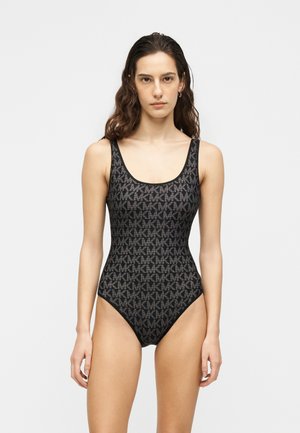 Femeie în picioare purtând un costum de baie negru fără mâneci cu model repetitiv gri al logo-ului „MK” pe un fundal alb simplu.