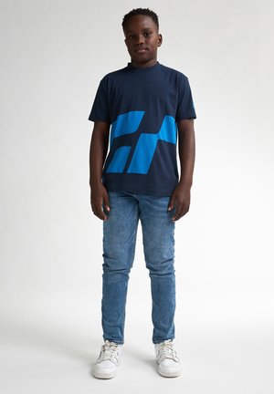 T-shirt in cotone navy con forme grafiche blu, abbinato a jeans in denim azzurro chiaro e sneaker bianche. Design semplice, maniche corte.