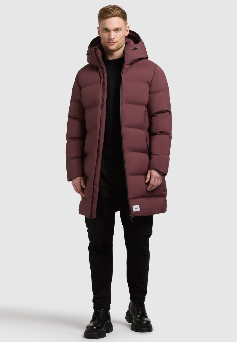 Maroon puffer coat with a hood, zip closure, and boxy design. Features a visible brand tag at the hem. Worn over a black sweater and pants.