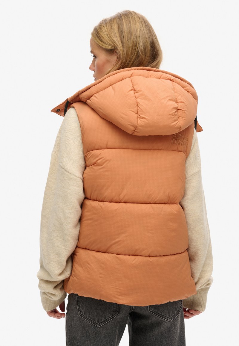 Kamizelka w kolorze przypalanej pomarańczy, z dużym kapturem, prążkowaną teksturą i detalem z logo. Nosi się ją na beżowym swetrze.