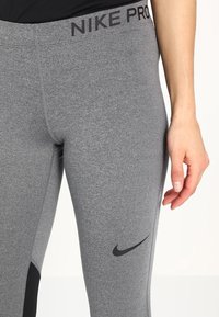 Les leggings Nike Pro gris présentent un design ajusté avec une taille large et un logo swoosh noir près de la cheville. Le tissu a une texture lisse.