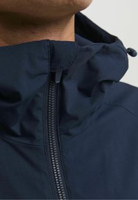 Giacca impermeabile blu navy con collo alto, cerniera frontale e coulisse regolabile sul cappuccio. Tessuto liscio e opaco senza motivi visibili.