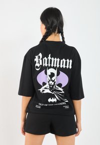 Black oversized t-shirt featuring a Batman graphic on the back. The design includes purple accents and bold white text. Paired with black shorts.