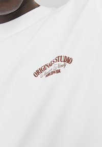 Camiseta blanca de algodón con cuello redondeado y mangas cortas, que presenta un logo impreso en marrón rojizo que dice "ORIGINALSSTUDIO Ninety Worldwide."
