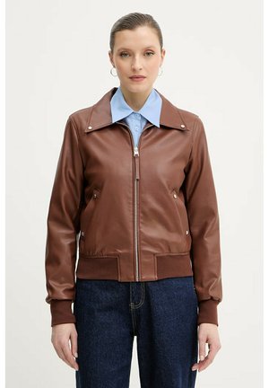 Donna che indossa una giacca marrone in pelle con zip sopra una camicia celeste con colletto e jeans di denim blu scuro, in piedi davanti a uno sfondo semplice.