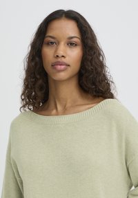 Pull vert clair en maille douce avec un large col côtelé et des épaules tombantes, présentant une texture lisse et une coupe décontractée.