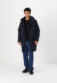 Parka bleu marine avec capuche, dotée de grandes poches avant et d'une fermeture éclair, portée sur une chemise noire et un jean bleu. Texture lisse, coupe décontractée.