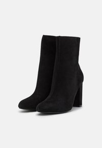 Černé suede kotníkové boty s špičatou špičkou, hladkou texturou a robustním vysokým podpatkem. Elegantní, minimalistický design bez viditelných kovových prvků.