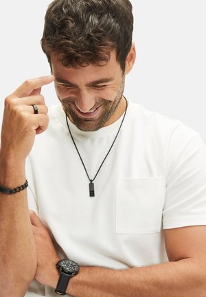 Zwarte hangers necklace aan een zwarte ketting. Witte T-shirt met zak. Zwarte armband en horloge om de pols. Man die glimlacht met kort, donker haar.