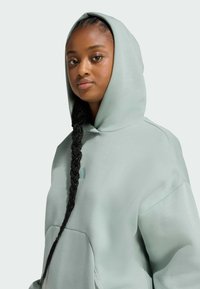 Mikina s kapucí v jasně zelené barvě s volným střihem, velkou kapsou na přední straně a malým logem na hrudi. Viditelné dlouhé pletené vlasy.