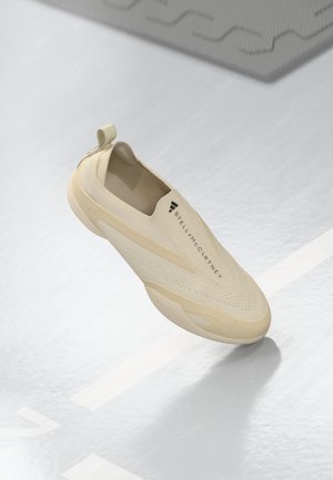 Beige slip-on sneaker with breathable knit upper and pull tab, featuring "Stella McCartney" branding, floating above glossy white floor.