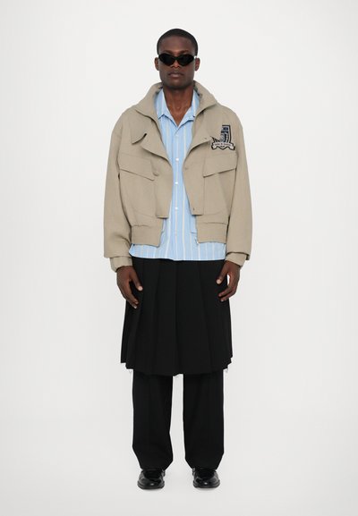 Beige Bouclé-Jacke mit großen Taschen, hellblau gestreiftes Hemd, schwarzer plissierter Rock und schwarze Schuhe; Model trägt eine Sonnenbrille.