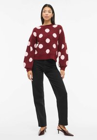 Suéter burdeos con grandes lunares rosas, tejido texturizado, cuello redondo y mangas globo. Combinado con pantalones negros y zuecos marrones con tacón.