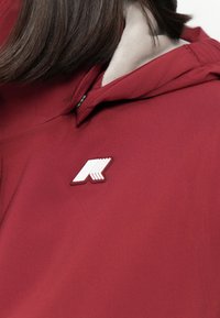 Rote Stoff-Hoodie mit einem auffälligen weißen Logo und einem schichtigen Design. Sichtbare glatte Textur und verstellbarer Kragen.