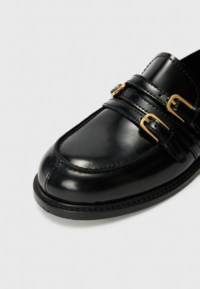 Mocasines de cuero negro con dos correas hebilladas, punta redondeada, acabado pulido y costuras visibles en la parte superior y la suela.