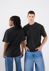 Tricouri negre, cu maneci scurte, de tip crew neck, cu o textură spălată, croială relaxată, purtate de două persoane care privesc în direcții diferite.