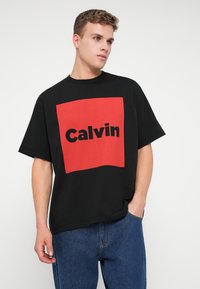 Crna majica s kratkim rukavima koja sadrži veliki crveni kvadrat s riječju "Calvin" ispisanom podebljanim crnim slovima. Opušteni kroj.