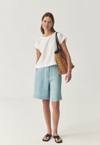 Witte, korte mouwen top gecombineerd met lichtblauwe shorts. Model houdt een geweven tan tas vast en draagt zwarte sandalen. Eenvoudig, casual ontwerp.