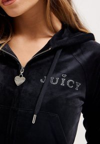 Felpa nera in velluto con zip, con scritta "JUICY" in strass, cappuccio, chiusura con zip e ciondolo a forma di cuore sulla cerniera.