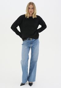 Zwarte gebreide trui met een lage schouderlijn, gecombineerd met lichtblauwe wijde jeans en puntige zwarte hakken, tegen een effen witte achtergrond.