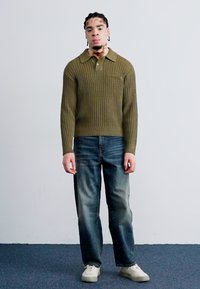 Olive green knitted sweater with a collar, paired with faded blue straight-leg jeans and beige sneakers, standing against a plain background.