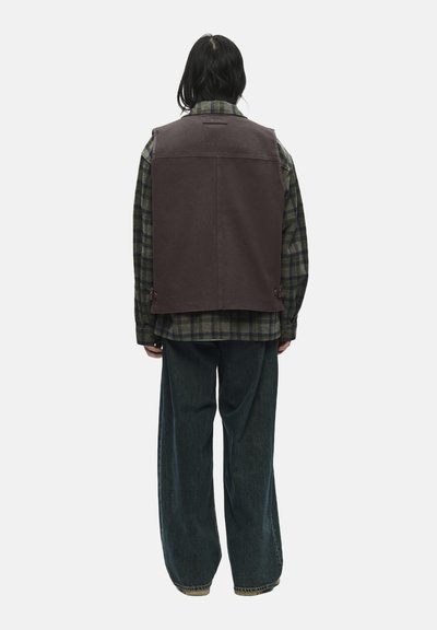 Vesta de țesătură maro purtată peste o cămașă cu carouri verzi, cu pantaloni din denim întunecat, lejeri. Prezintă o croială dreaptă și detalii minime pe spate.
