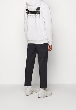 Person wearing white hooded sweatshirt with black text, black cropped trousers, white socks, and chunky white sneakers standing against white background.