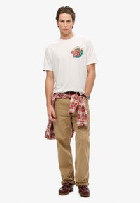 Camiseta blanca con un gráfico colorido de "SUPER DRY", pantalones caqui y una camisa de franela a cuadros rojos atada a la cintura; zapatos marrones con cordones.