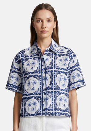 Donna che indossa una camicia blu e bianca a maniche corte con bottoni, decorata con motivo di palme e paesaggi, abbinata a pantaloni bianchi.