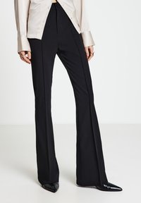 Schwarze Schlaghosen aus glattem Stoff mit hoher Taille und maßgeschneiderter Passform. Kombiniert mit spitzen schwarzen Schuhen und einer cremefarbenen Bluse.