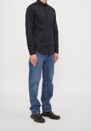Hombre de pie vestido con una camisa negra abotonada, jeans azules y zapatos negros de punta cuadrada contra un fondo claro y liso.