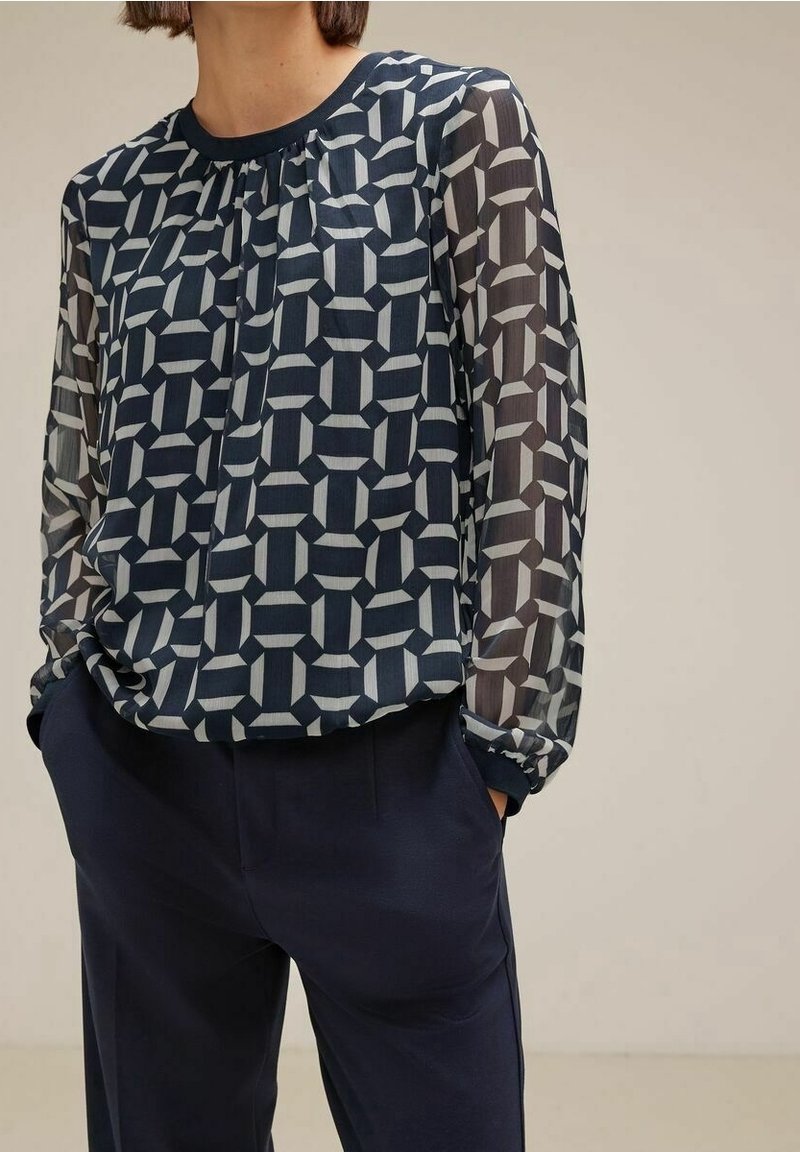 Femme portant un chemisier transparent à manches longues bleu marine à motifs, associé à un pantalon taille haute bleu marine foncé, les mains dans les poches.