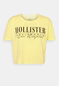 Žluté zkrácené tričko z bavlny s kulatým výstřihem. Obsahuje tištěný text tmavě hnědou barvou: "HOLLISTER CALIFORNIA Los Angeles."