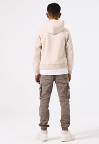 Felpa beige con un grande cappuccio, abbinata a pantaloni cargo grigi con tasche laterali e polsini elasticizzati, completata da sneakers bianche.