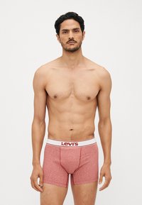 Crvene bokserice s mekim, teksturiranim materijalom i bijelim pojasom na kojem se nalazi "Levi's" logo u crvenoj boji. Duljina do sredine bedra i uski dizajn.