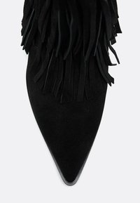 Scarpa nera in suede con tacco alto, caratterizzata da dettagli in frange alla caviglia e una punta appuntita. Texture liscia con una suola sottile.