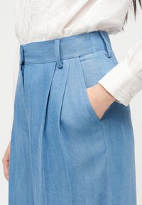 Γυναίκα που φοράει ανοιχτό μπλε πλισέ παντελόνι με ψηλή μέση και χέρι στην τσέπη, σε συνδυασμό με άσπρο πουκάμισο με δαντελένια μανσέτα.