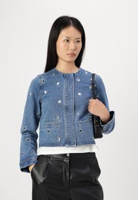 Jeans jakna z značilnimi srebrnimi luknjicami, zaobljenim izrezom, skrajšano obliko in dvema sprednjima žepoma, v kombinaciji z belo majico in črnimi hlačami.