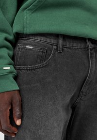 Jeans in denim grigio con una texture sottile, dotati di tasche anteriori e di un bottone in metallo. Una felpa verde parzialmente visibile nella parte superiore.
