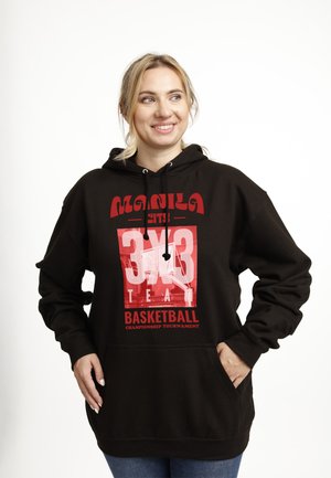 Uśmiechnięta kobieta w czarnej bluzie z kapturem z czerwonym napisem „Manila City 3x3 Team Basketball Championship Tournament”, stoi z ręką w kieszeni.