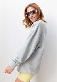 Grauer, übergroßer Pullover mit langen Ärmeln, gerippten Bündchen und rundem Ausschnitt. Kombiniert mit gelben, gerippten Radlerhosen und großen Sonnenbrillen.