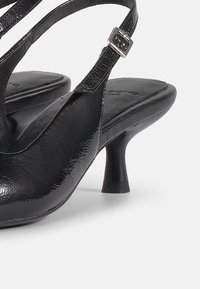 Chaussure slingback en cuir noir avec un petit talon courbé et une bride réglable à boucle argentée sur fond blanc.