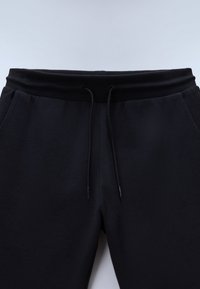 Pantalon de survêtement noir avec ceinture côtelée et cordons noirs, doté de poches latérales, présenté sur un fond clair uni.